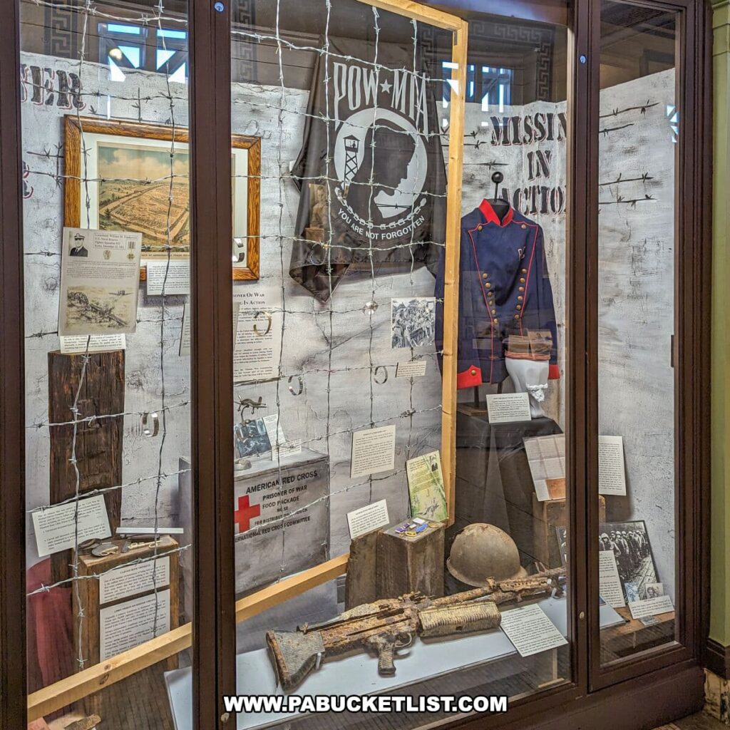 Exhibit honoring prisoners of war and those missing in action at the Soldiers and Sailors Memorial Hall and Museum in Pittsburgh, PA. The display features a POW/MIA flag, military artifacts, photographs, and personal items behind a glass case. The backdrop is a wall with barbed wire graphics and information panels, including a Red Cross food package, creating a somber reflection on the sacrifices of service members.