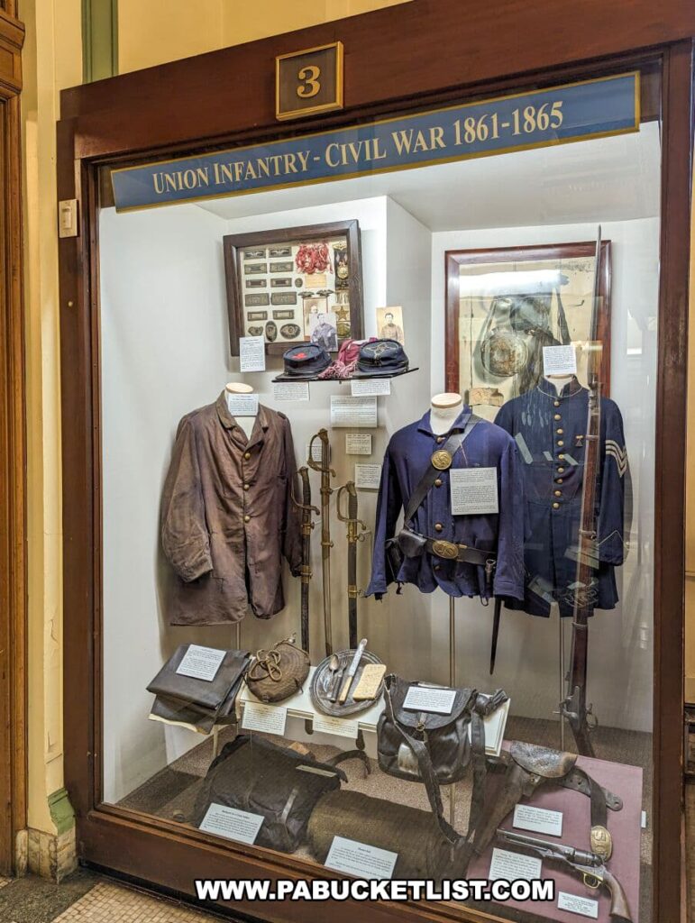 Exhibit at the Soldiers and Sailors Memorial Hall and Museum in Pittsburgh, PA, showcasing Union Infantry uniforms and equipment from the Civil War era, 1861-1865. The display includes two mannequins dressed in period uniforms, a selection of hats, military accoutrements, a rifle, and informational placards providing historical context on the items presented.