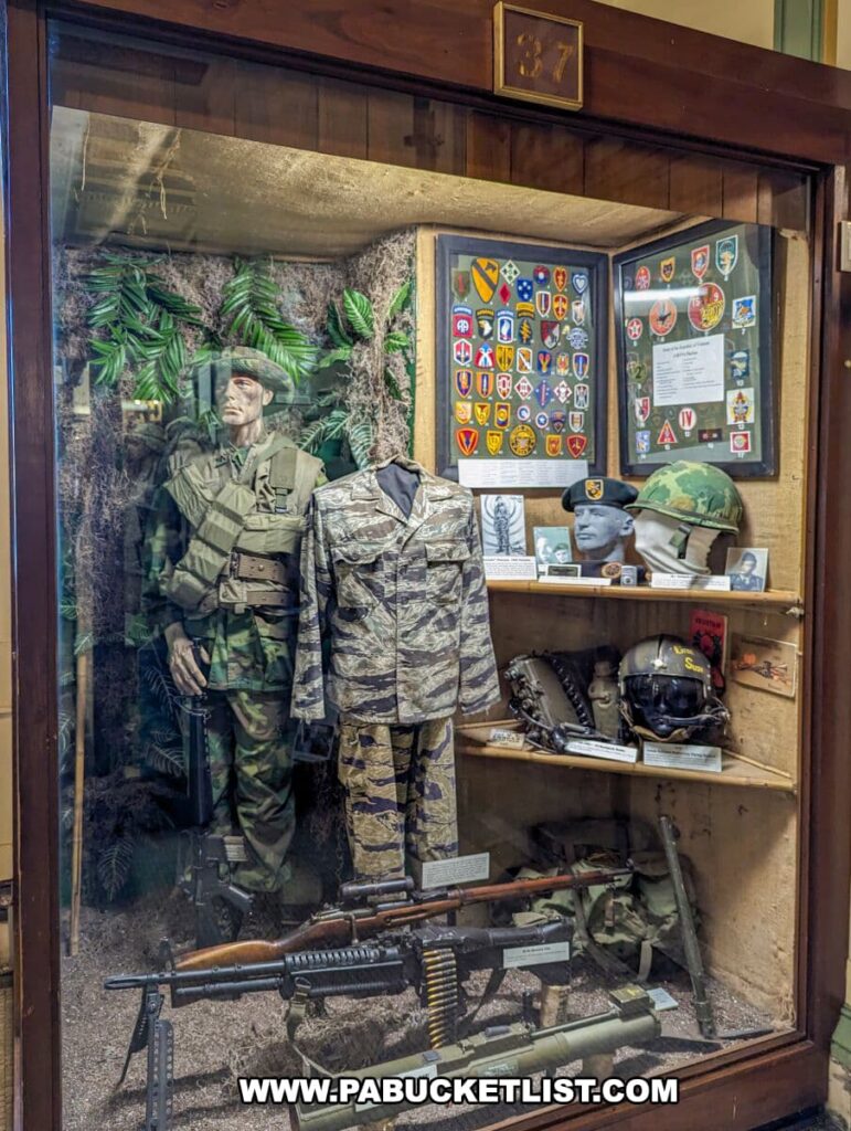 Vietnam War memorabilia exhibit at the Soldiers and Sailors Memorial Hall and Museum in Pittsburgh, PA. The display features a mannequin dressed in a camouflage uniform, military patches, a collection of helmets, and a variety of infantry weapons. The backdrop includes lush green foliage, creating an immersive environment reminiscent of the Vietnamese jungle where American soldiers were deployed.