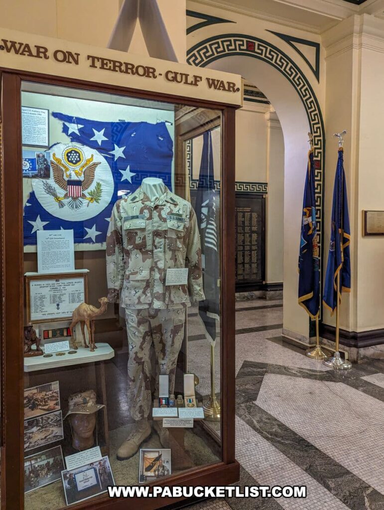 The 'War on Terror - Gulf War' exhibit at the Soldiers and Sailors Memorial Hall and Museum in Pittsburgh, PA, showcasing a mannequin clad in desert camouflage military uniform, a flag with the Great Seal of the United States, and various military artifacts and personal items, including a camel figurine. The display is set against the backdrop of the museum's elegant interior architecture.