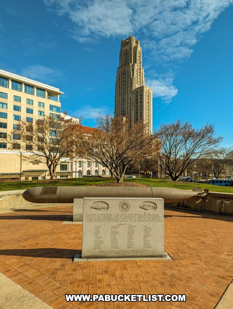 Outdoor memorial at the Soldiers and Sailors Memorial Hall and Museum in Pittsburgh, PA, featuring a large naval torpedo on display. In the background, the iconic Cathedral of Learning towers over the scene. A commemorative plaque in the foreground lists the names of submariners lost at sea, with a brick pavement leading up to the monument under a clear blue sky.