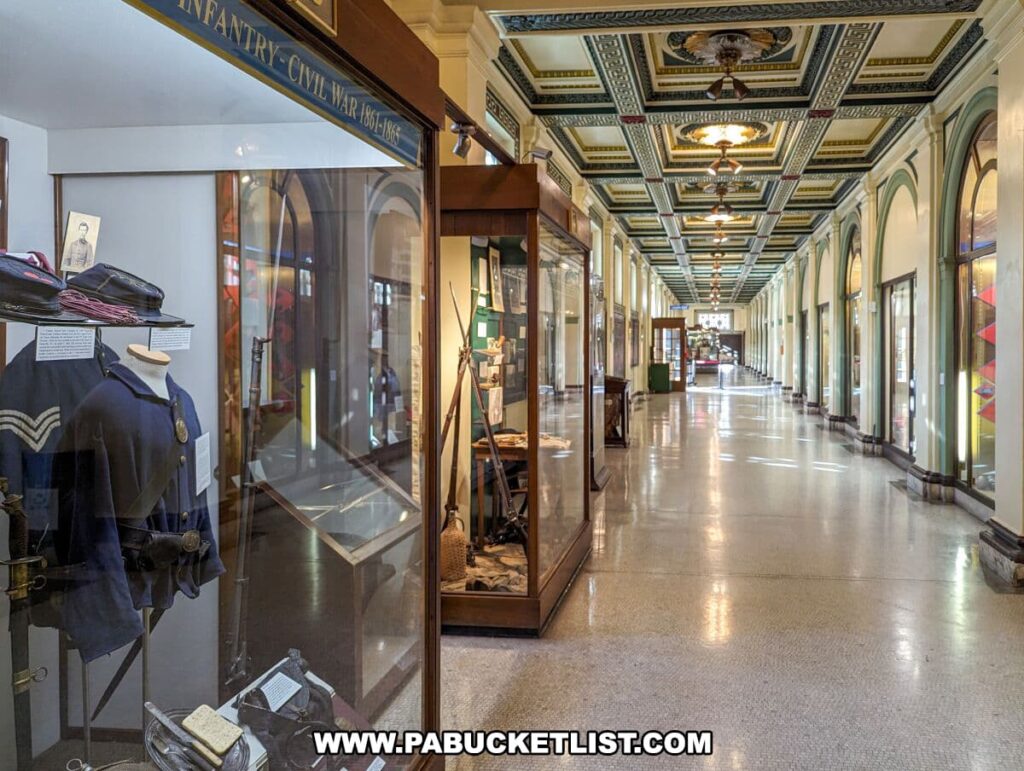 A long, elegant corridor inside the Soldiers and Sailors Memorial Hall and Museum in Pittsburgh, PA, lined with glass cases displaying Civil War memorabilia. The hall is illuminated by period light fixtures, with detailed ceiling decorations and terrazzo flooring that reflect the museum's historic charm.