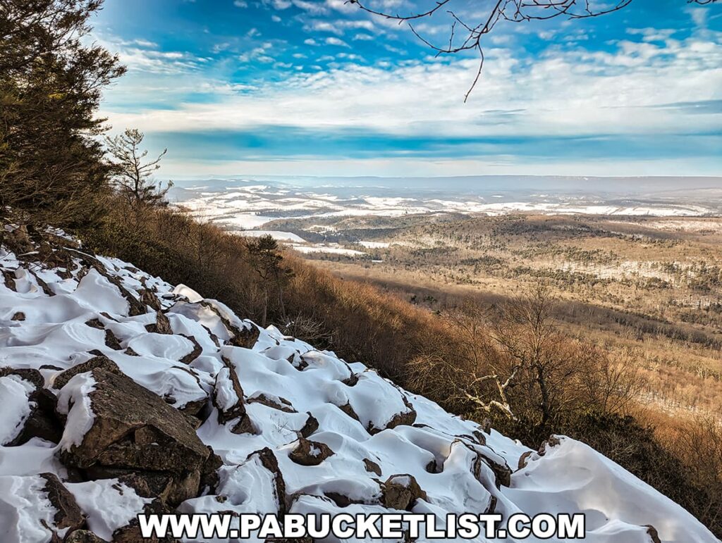 A scenic view from the Stone Valley Vista on the Standing Stone Trail in Huntingdon County, PA, featuring a snowy slope with jagged rocks protruding through the white blanket. Evergreen trees to the left and leafless branches to the right frame the vista, which overlooks a vast valley with patches of snow, forested areas, and distant hills under a partly cloudy sky with soft blue tones.