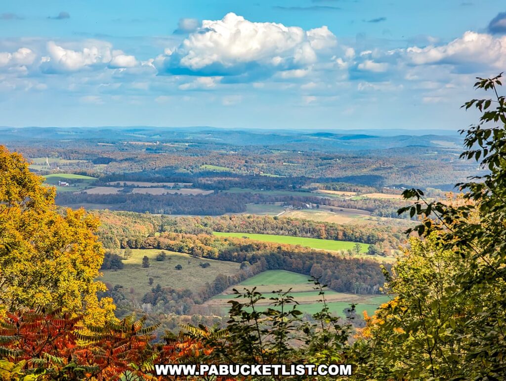 A panoramic view from Mount Pisgah County Park in Bradford County, Pennsylvania. The landscape is a tapestry of autumn colors with patches of farmland nestled among rolling hills. The foreground features trees ablaze with oranges and yellows, while the background extends into distant blue-green hills under a sky dotted with fluffy white clouds. The scene captures the serene beauty of rural Pennsylvania in the fall.