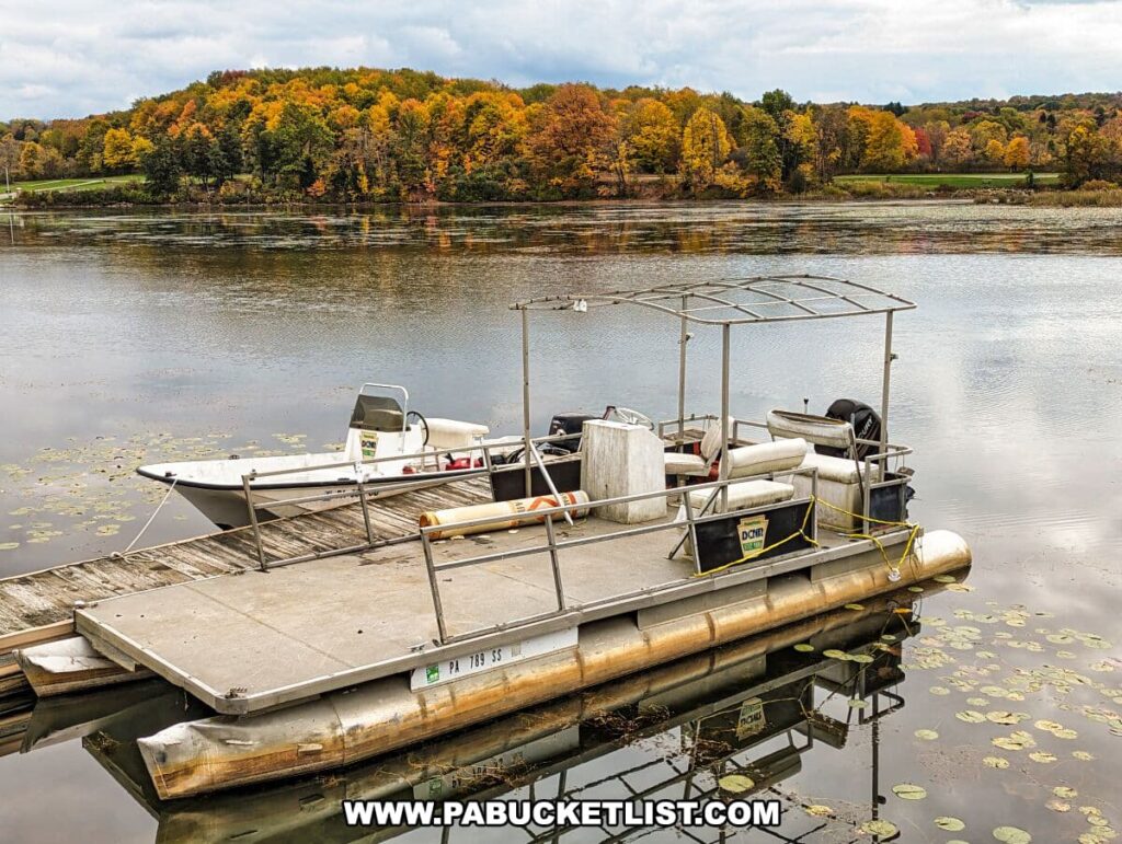 A floating dock at Yellow Creek State Park in Indiana County, Pennsylvania, with two pontoon boats moored to it. One of the boats is equipped with a canopy and appears ready for use, while the other is partially covered with a tarp, possibly for maintenance. The water is calm with a scattering of lily pads, reflecting the cloudy sky. The background showcases a vibrant display of autumn foliage with trees in shades of yellow, orange, and red. A label on the boats indicates they are property of the Department of Conservation and Natural Resources (DCNR) of Pennsylvania.