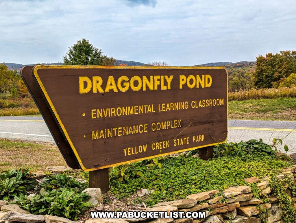 A wooden sign at Yellow Creek State Park in Indiana County, Pennsylvania, reading "DRAGONFLY POND" in bold yellow letters against a dark background. Below, the sign lists "Environmental Learning Classroom" and "Maintenance Complex," followed by "Yellow Creek State Park," indicating the amenities and the park's name. The sign is positioned at the side of a road, with a stone wall and greenery at its base. In the background, a hilly landscape with trees beginning to show autumn colors can be seen under an overcast sky.