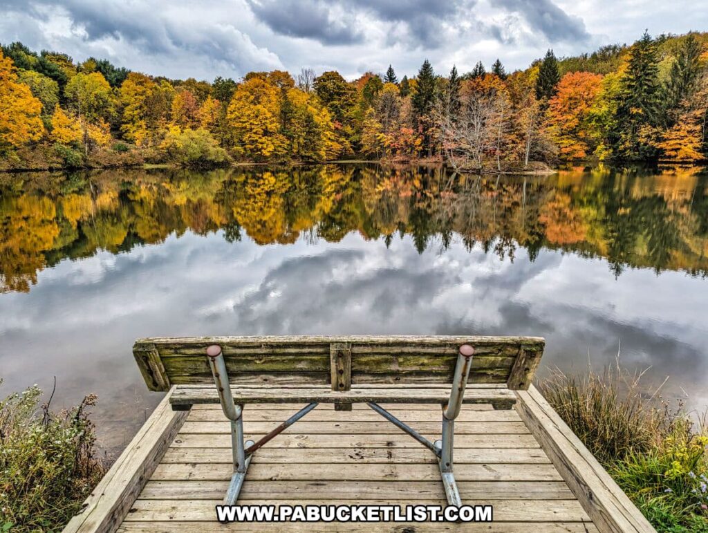 A serene autumnal scene at Yellow Creek State Park in Indiana County, Pennsylvania. The image captures a wooden dock overlooking Dragonfly Pond, with the calm waters reflecting the vibrant fall foliage. The trees display a palette of warm colors ranging from yellow to orange and red, contrasting against the evergreens. The sky is overcast with dynamic cloud formations, and the peacefulness of the scene is palpable.
