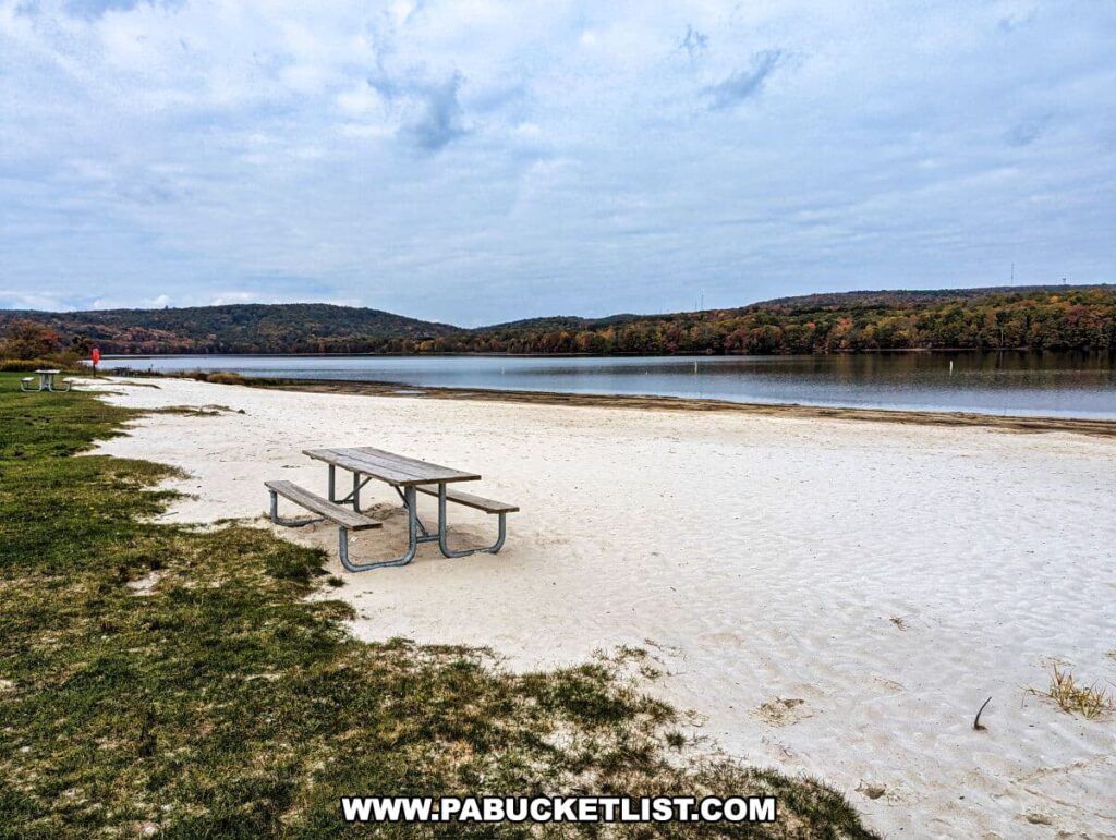 A tranquil lakeside scene at Yellow Creek State Park in Indiana County, Pennsylvania. It shows a sandy beach in the foreground with a single picnic table and bench, inviting a peaceful rest with a view. The lake stretches out toward the horizon, bordered by a lush, hilly landscape adorned with a mosaic of autumn colors. A lifeguard chair stands empty, signaling the off-season. Overhead, the sky is partly cloudy, suggesting a cool autumn day.