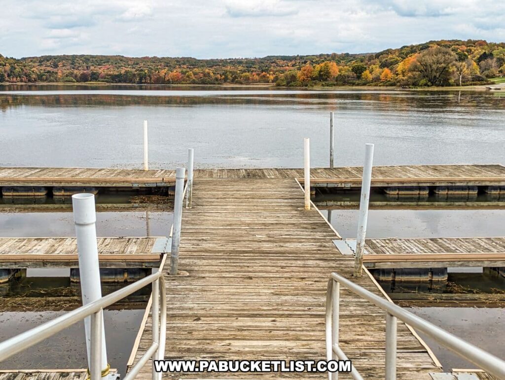 A wooden boat dock at Yellow Creek State Park in Indiana County, Pennsylvania. The perspective is from the shore, looking out toward the water. The dock extends into the lake, flanked by metal railings and equipped with vertical posts for mooring boats. The water is still, mirroring the cloudy sky and the vibrant fall foliage on the distant hills. The trees display an array of colors, including yellow, orange, and red, indicative of the autumn season.