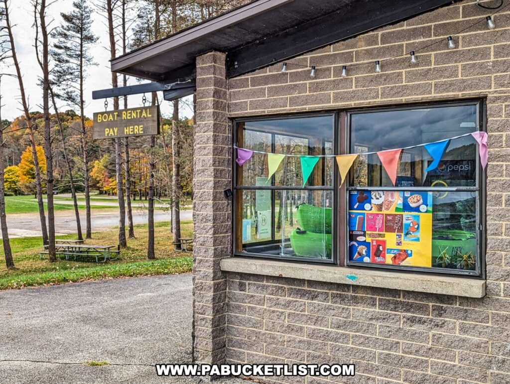 The exterior of a boat rental facility at Yellow Creek State Park in Indiana County, Pennsylvania. The building has a brick facade with a large window displaying colorful ice cream advertisements and a Pepsi logo. A string of multicolored triangular flags hangs in the window, adding a festive touch. Above the window, a sign reads "BOAT RENTAL PAY HERE," indicating the service provided. The surrounding area is visible through the reflection on the window and includes trees with autumn foliage and a picnic area with tables.