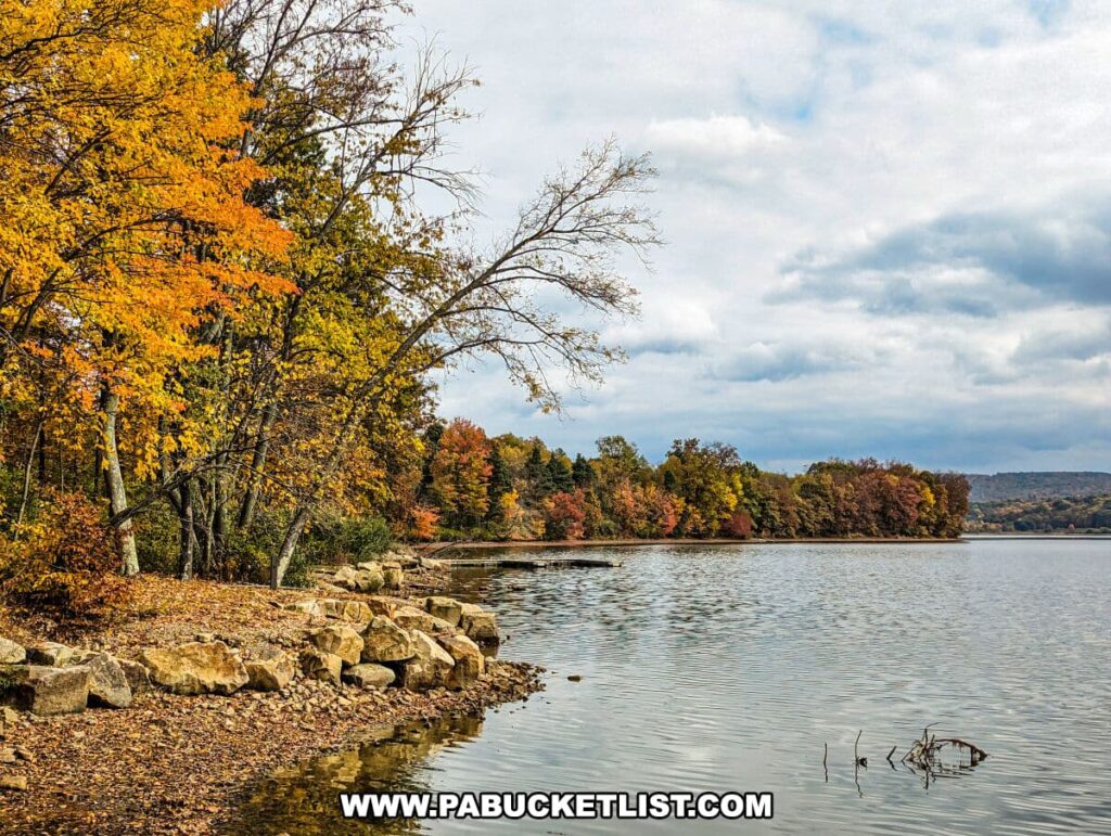 A scenic view along the north shore of Yellow Creek State Park in Indiana County, Pennsylvania. The lake's edge is lined with a rocky embankment and a tree with bright yellow autumn leaves. In the background, the opposite shore is covered with a forest showcasing a colorful mix of fall foliage in shades of red, orange, and green. The water is calm, reflecting the cloudy sky and the autumnal trees.