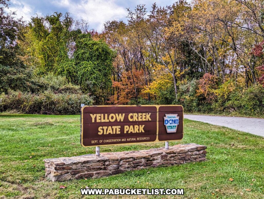 The entrance sign for Yellow Creek State Park in Indiana County, Pennsylvania. The sign is brown with white lettering stating "YELLOW CREEK STATE PARK" and includes the Pennsylvania Department of Conservation and Natural Resources (DCNR) logo. The sign is mounted on a stone base and set against a backdrop of lush trees in various autumnal colors. A grassy area surrounds the sign, and a paved road can be seen curving beside it.