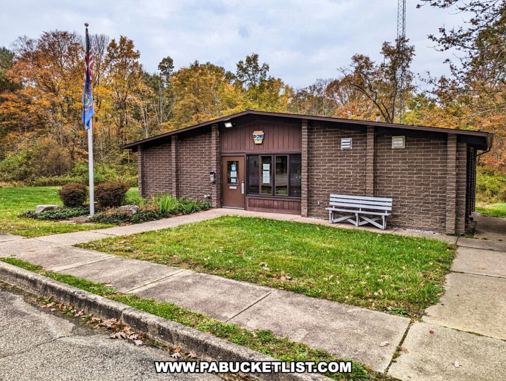 The park office at Yellow Creek State Park in Indiana County, Pennsylvania. The small, one-story building has a dark brown brick exterior with a lighter brown trim around the windows and doors. A white wooden bench sits in front of the building. The American and Pennsylvania state flags are flying on a flagpole to the left of the entrance. The office is set against a backdrop of trees displaying autumn foliage in shades of yellow and orange.