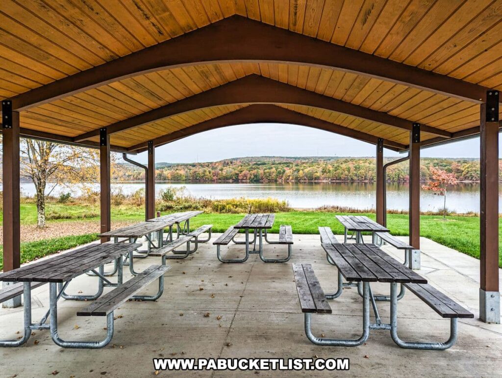 A picnic pavilion at Yellow Creek State Park in Indiana County, Pennsylvania. The pavilion has a wooden gabled roof supported by metal beams and houses several picnic tables made of wood with metal frames. The pavilion overlooks a scenic view of a lake with trees displaying autumn colors on the far shore. The ground outside the pavilion is covered with grass and fallen leaves, indicative of the fall season. The sky is overcast, and the peaceful lake reflects the grey clouds above.