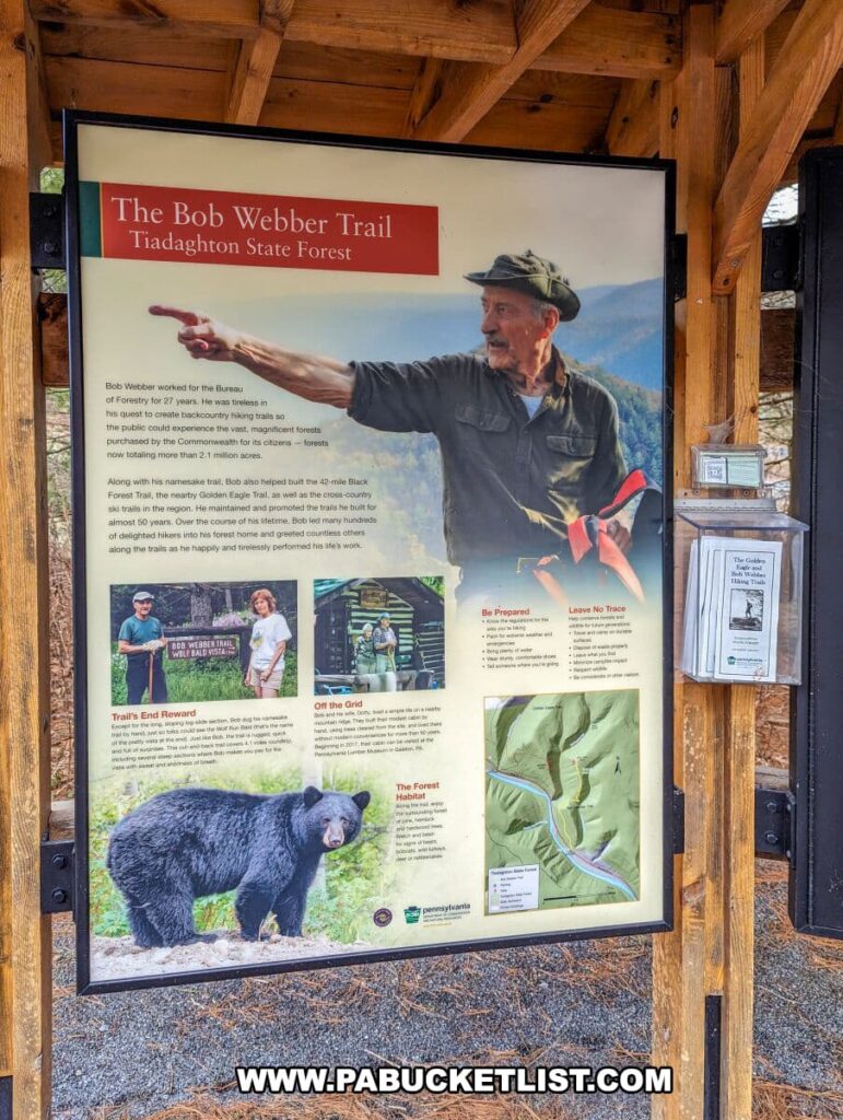 An informational sign about The Bob Webber Trail in Tiadaghton State Forest, displayed within a wooden shelter. The sign features a photograph of Bob Webber pointing towards a scenic view, a map of the trail, and several smaller images, including hikers and a black bear. Text on the sign provides background on Bob Webber and trail advice like "Be Prepared" and "Leave No Trace." The sign emphasizes the natural beauty of the area, the enjoyment of hiking, and the importance of conservation.