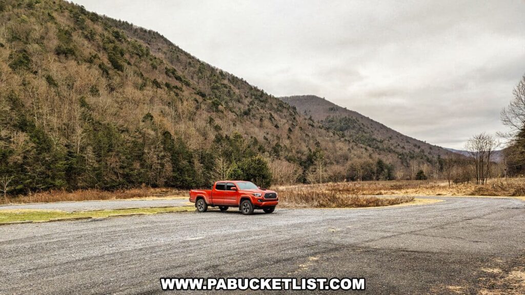An orange pickup truck parked on a gravel lot in the Tiadaghton State Forest, with the Bob Webber Trail nearby. The forested Pennsylvania mountains rise in the background, with leafless trees on the lower slopes indicating either autumn or winter. The sky is overcast, and the open field next to the parking area is filled with tall, dry grasses, contributing to a serene natural landscape.