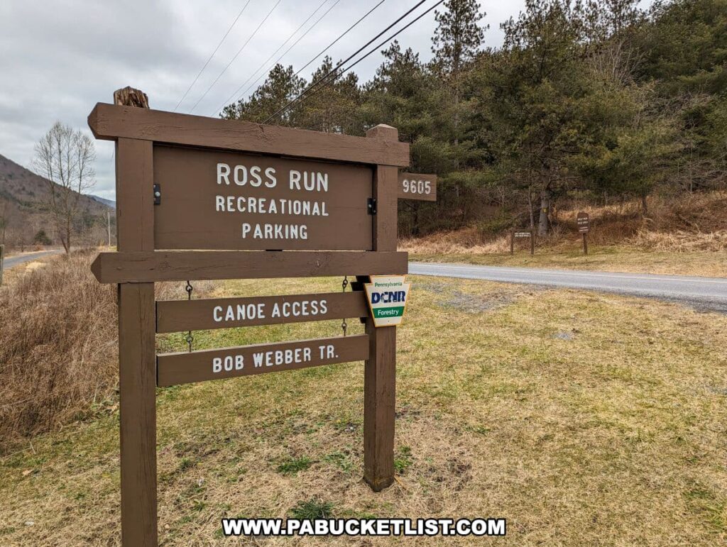 A wooden signpost at the Ross Run Recreational Parking area, indicating the starting point for the Bob Webber Trail and canoe access in Tiadaghton State Forest, Lycoming County, Pennsylvania. The signpost, showing "Ross Run Recreational Parking" and "Canoe Access Bob Webber Tr.," stands prominently on the grass verge by the roadside, with the forest and a mountainous backdrop in the distance. An additional sign for Pennsylvania DCNR Forestry is attached to the post, suggesting the area is managed for recreational activities.