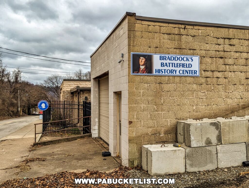 Exterior view of Braddock's Battlefield History Center near Pittsburgh, featuring a simple, single-story building with cinder block walls. On the side of the building, a large sign with an image of General Braddock and the text "Braddock's Battlefield History Center" is displayed. In the foreground, there is a blue historic marker sign with a circular emblem. The center is surrounded by a bare tree landscape under an overcast sky.