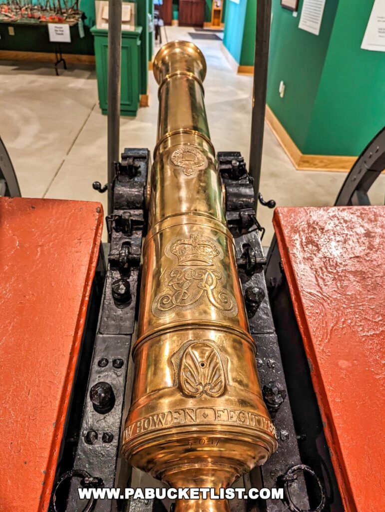 A close-up view of a shiny brass cannon on display at Braddock's Battlefield History Center near Pittsburgh, showcasing detailed engravings, including a royal crest and a fleur-de-lis. The cannon is mounted on a black carriage with red trim, hinting at its historical British military use. The backdrop features the green walls of the museum interior.