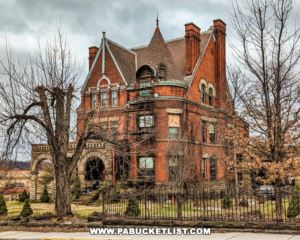 A photograph of the former Charles Schwab mansion, near Braddock's Battlefield History Center. The Victorian-era mansion features red brickwork, multiple gables, a prominent conical-roofed turret, and stone arches above the entrance. The building is enclosed by a wrought iron fence and is framed by bare trees, under a cloudy sky.
