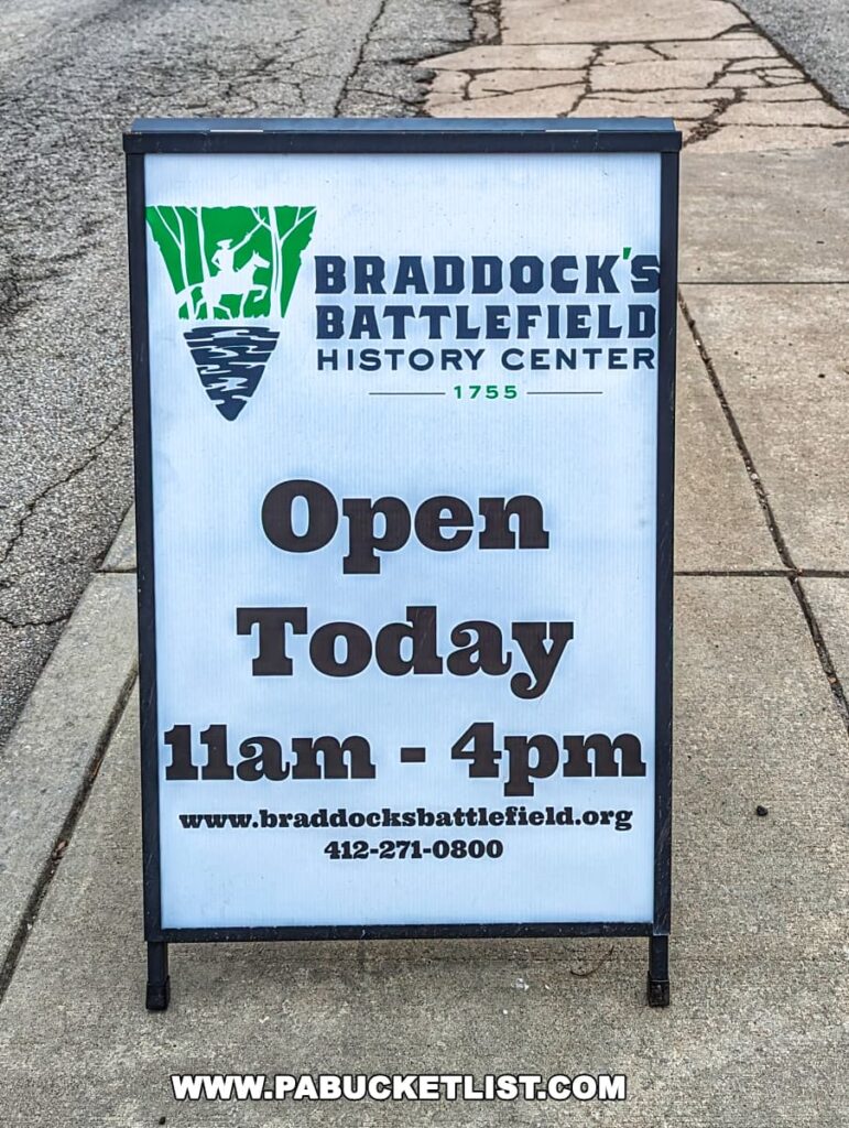 Signage for Braddock's Battlefield History Center indicating it is open today from 11 am to 4 pm. The sign features the center's logo, a green silhouette of a soldier on a white and green shield with the year 1755, and includes the website URL and a contact phone number. The sign is placed on a sidewalk with a cracked pavement background.