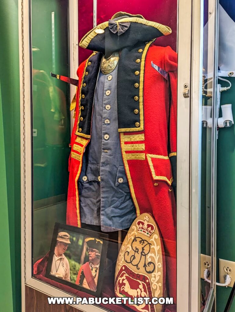 A display case at Braddock's Battlefield History Center showcasing a replica of a British military uniform from the mid-18th century. The uniform includes a red coat with blue facings, gold braid, and brass buttons, a black tricorne hat with gold trim, and a white waistcoat. Below the uniform, a framed photograph depicts reenactors in period attire, and a decorative drum with historical motifs is also visible.