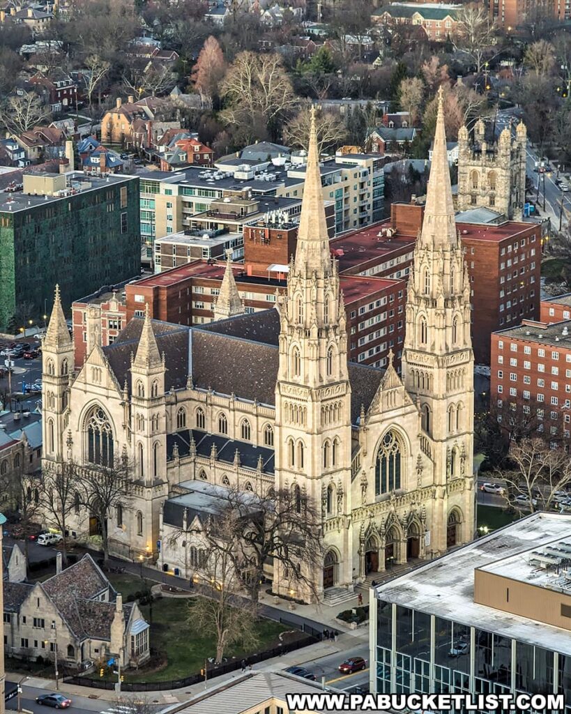 A view of Saint Paul Cathedral in Pittsburgh, Pennsylvania from the Cathedral of Learning at Pitt. The cathedral displays intricate Gothic Revival architecture with pointed arches and towering spires. The building is nestled among the urban landscape of the city, surrounded by residential buildings, trees, and streets with cars. The foreground shows a modern building with a glass facade that contrasts with the historical and ornate design of the cathedral. The photo captures the blending of architectural styles in an urban setting.