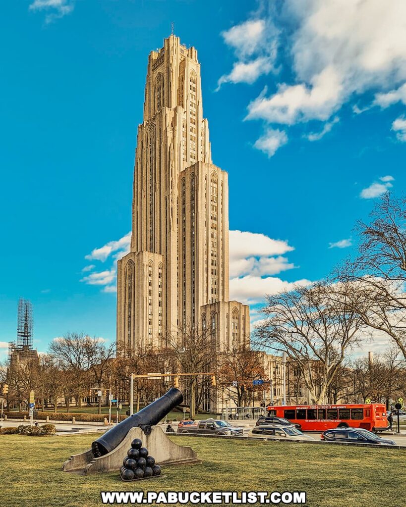 An image of the Cathedral of Learning at the University of Pittsburgh, a tall, Gothic Revival skyscraper under a blue sky with scattered clouds. In the foreground is a large, historic cannon displayed on a concrete base with cannonballs stacked at its base, situated on a well-manicured lawn. Traffic is visible on the adjacent road, including a red bus among the cars. The scene suggests a clear day with the bare trees indicating either late fall or winter.
