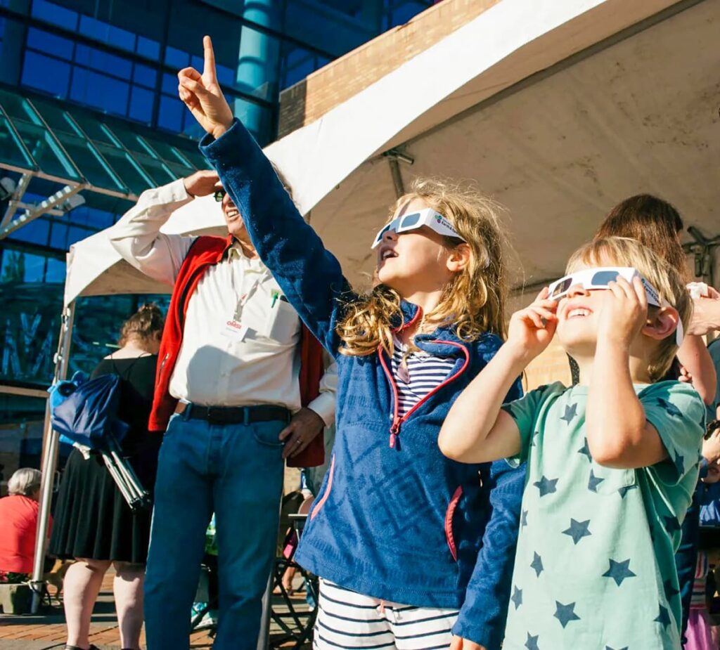 A group of excited children and adults wearing protective eclipse glasses looking up at the sky during the 2024 total solar eclipse, with one man pointing upwards, indicating the event. They are outdoors, gathered in a public viewing area with a modern building in the background, under clear blue skies.