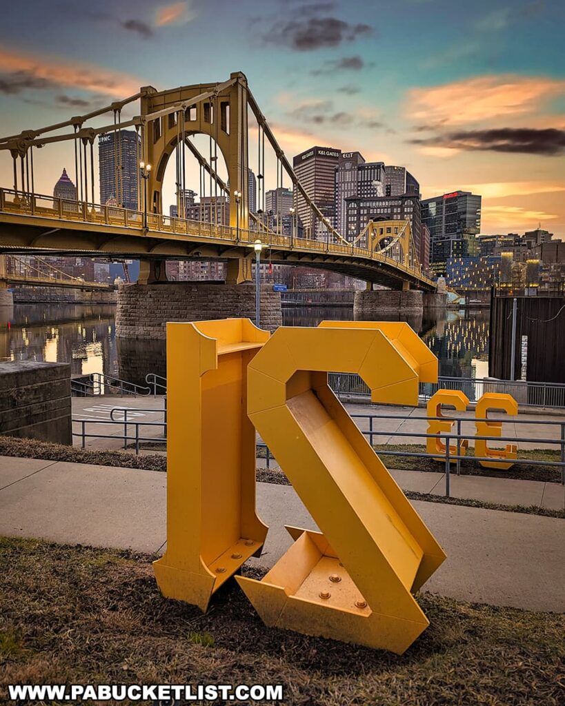 A photograph captured at dusk featuring the Roberto Clemente Bridge in Pittsburgh, Pennsylvania. In the foreground, a large yellow numeral '21' sculpture stands prominently, with a smaller '33' sculpture in the background. The bridge, also painted yellow, spans the Allegheny River and leads to the city skyline, which is lit up against the fading light of the sky showing hues of pink, blue, and orange. The water reflects the bridge and city lights, adding to the serene evening atmosphere.