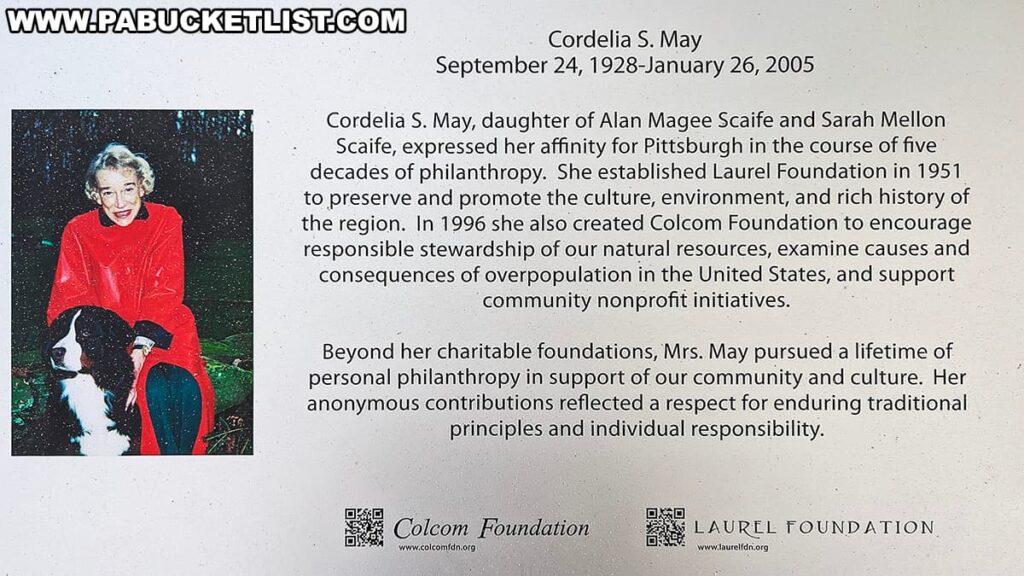A commemorative plaque dedicated to Cordelia S. May, featuring a brief biography and her contributions to philanthropy. There's a photograph of Cordelia May smiling, dressed in a red coat, kneeling beside a black and white dog. The text mentions her establishment of the Laurel Foundation in 1951 and the Colcom Foundation in 1996 to support culture, environment, and community initiatives. The plaque highlights her lifetime of personal philanthropy and anonymous contributions that reflect her respect for traditional principles and individual responsibility. The logos for Colcom Foundation and Laurel Foundation, along with their websites, are displayed at the bottom.
