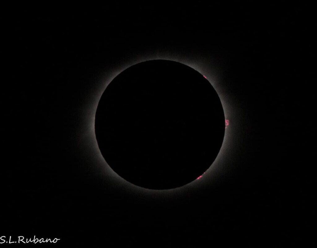 A striking image of the 2024 total solar eclipse as seen from Pennsylvania, showcasing the phenomenon where the Moon completely covers the Sun, leaving only the glowing solar corona visible. Small pinkish solar prominences can be seen around the edge of the silhouette. The background is predominantly dark, highlighting the eclipse's dramatic contrast.