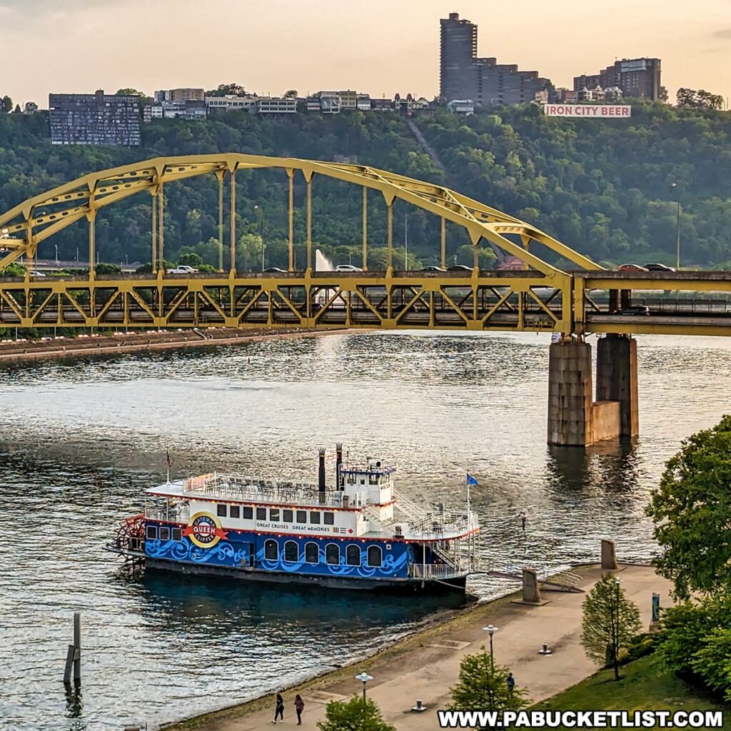 An image capturing the Gateway Clipper, a blue and white paddle steamer, on the Allegheny River in Pittsburgh, Pennsylvania. The boat is in the foreground, with the city's iconic yellow Fort Duquesne Bridge arching overhead. In the background, the green hills of Mount Washington and residential buildings are visible, along with the "IRON CITY BEER" sign. The evening light gives a warm hue to the scene, with a clear sky above and a few people visible walking along the riverbank.
