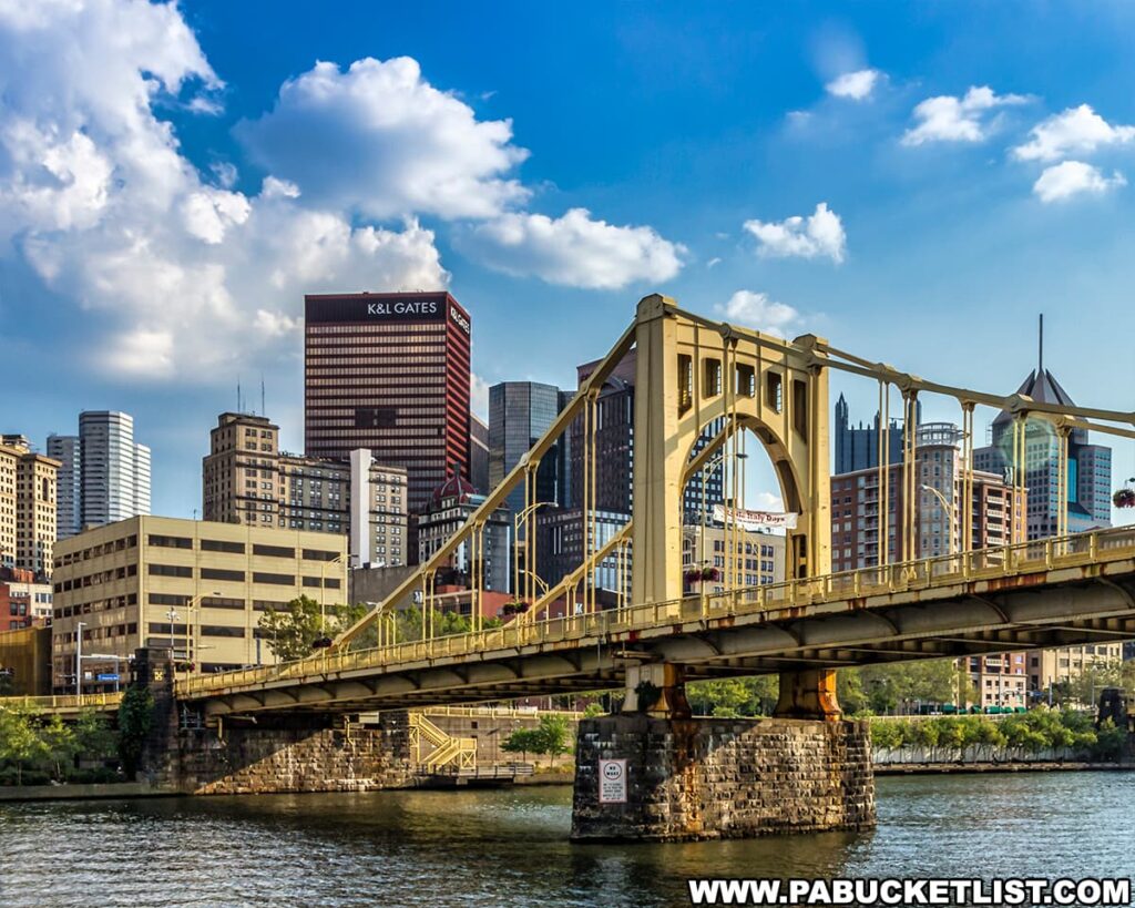 A view of the Pittsburgh, Pennsylvania skyline featuring the iconic yellow Roberto Clemente Bridge in the foreground. The K&L Gates Center is prominent among the array of downtown buildings. The Allegheny River is in the foreground, reflecting the bridge and parts of the cityscape. The sky is blue with scattered clouds on a bright, sunny day.