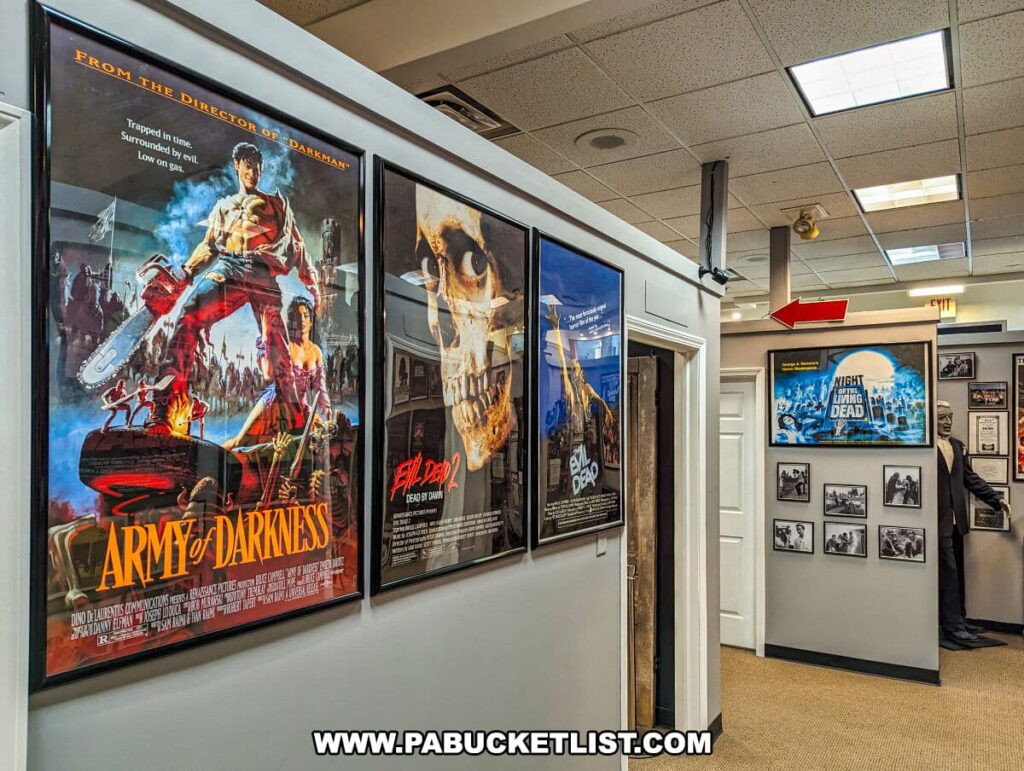 Interior of the Living Dead Museum showcasing framed movie posters on white walls. On the left, a prominent poster of 'Army of Darkness' features a heroic figure with a chainsaw arm. Adjacent is the 'Evil Dead II' poster with a skull. Towards the back, the 'Night of the Living Dead' is displayed above black and white photos. The museum has ceiling tiles, fluorescent lighting, and a figure can be partially seen to the right, suggesting more exhibits.