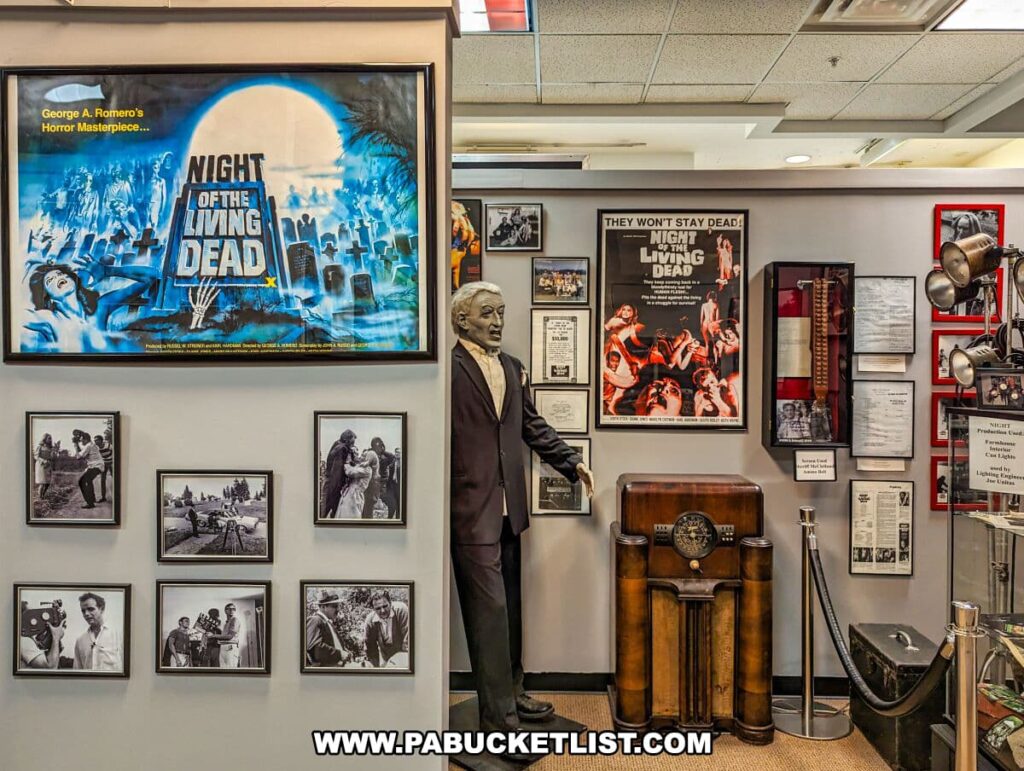 An exhibit inside the Living Dead Museum featuring memorabilia from George A. Romero's 'Night of the Living Dead'. On the left, a large poster with a vivid blue and yellow design is displayed above a series of black and white photos from the movie's production. To the right, a life-size statue of Romero stands beside a wall with another movie poster and a display case containing various historical documents and props, including an antique radio. The exhibit captures the essence of the classic film and honors the legacy of its creator.