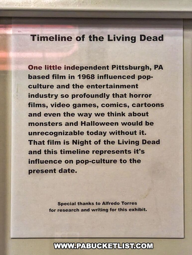 An information panel at the Living Dead Museum titled 'Timeline of the Living Dead', acknowledging the significant impact of the 1968 Pittsburgh-based film 'Night of the Living Dead' on popular culture and the entertainment industry. The text credits the film with profoundly influencing horror films, video games, comics, cartoons, and the general perception of monsters and Halloween. A note at the bottom expresses gratitude to Alfredo Torres for research and writing for the exhibit, highlighting the collaborative effort behind the museum's educational content.