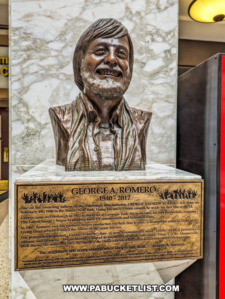 A bronze bust of filmmaker George A. Romero, displayed at the Living Dead Museum. Romero is depicted with a cheerful expression, wearing a jacket and shirt. The bust is mounted on a pedestal featuring a plaque with a detailed biography and accolades, including his life span from 1940 to 2017, his contributions to the Pittsburgh film industry, and his pioneering work on 'Night of the Living Dead' and 'Dawn of the Dead'. The backdrop has a marble texture, with a partial view of the mall's interior, including a column and a lit lamp, indicating the museum's location inside the Monroeville Mall.