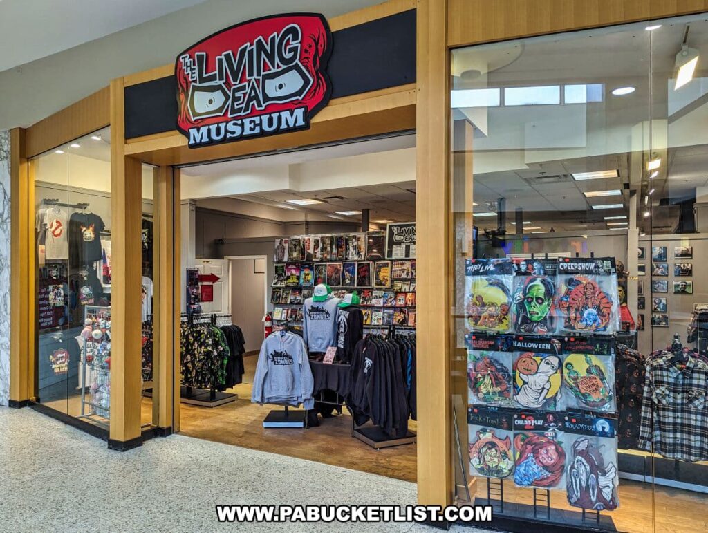The entrance to the Living Dead Museum located in the Monroeville Mall. The museum's bold sign features zombie eyes and is mounted above the doorway. The glass storefront displays an array of merchandise, including t-shirts and posters with horror and zombie-themed graphics. Featured items showcase iconic horror movie imagery, such as 'Creepshow' and 'Halloween'. The store is situated on a tiled floor within the mall, inviting fans of the genre and George Romero's work to explore its content.