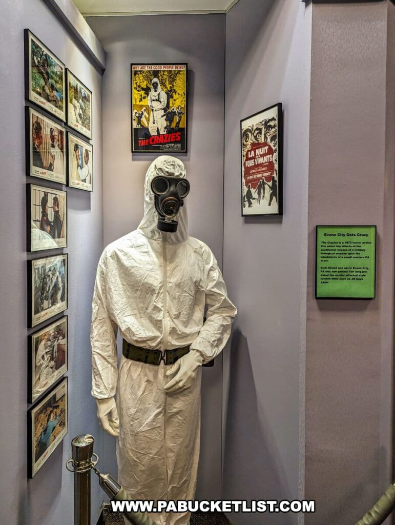 An exhibit at the Living Dead Museum dedicated to 'The Crazies' film, featuring a mannequin dressed in a white hazmat suit with a gas mask. The display is surrounded by film stills and posters, including the prominent yellow poster of 'The Crazies' with the tagline 'Why are the good people dying?'. To the right, a European film poster adds international context. An informational plaque titled 'Evans City Gets Crazy' details the film's local shooting. This corner of the museum immerses visitors in the chilling atmosphere of the movie.