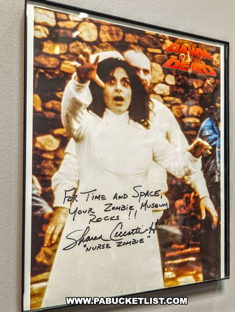 A signed photo on display at the Living Dead Museum featuring Sharon Ceccatti-Hill, known as the 'Nurse Zombie' from 'Dawn of the Dead'. She is depicted in her costume, reaching out in a classic zombie pose. The inscription reads 'For Time and Space, Your Zombie Museum Rocks!!' above her signature and character name, expressing appreciation for the museum. The photograph is a tangible piece of the film's history, connecting fans to the iconic characters of the series.