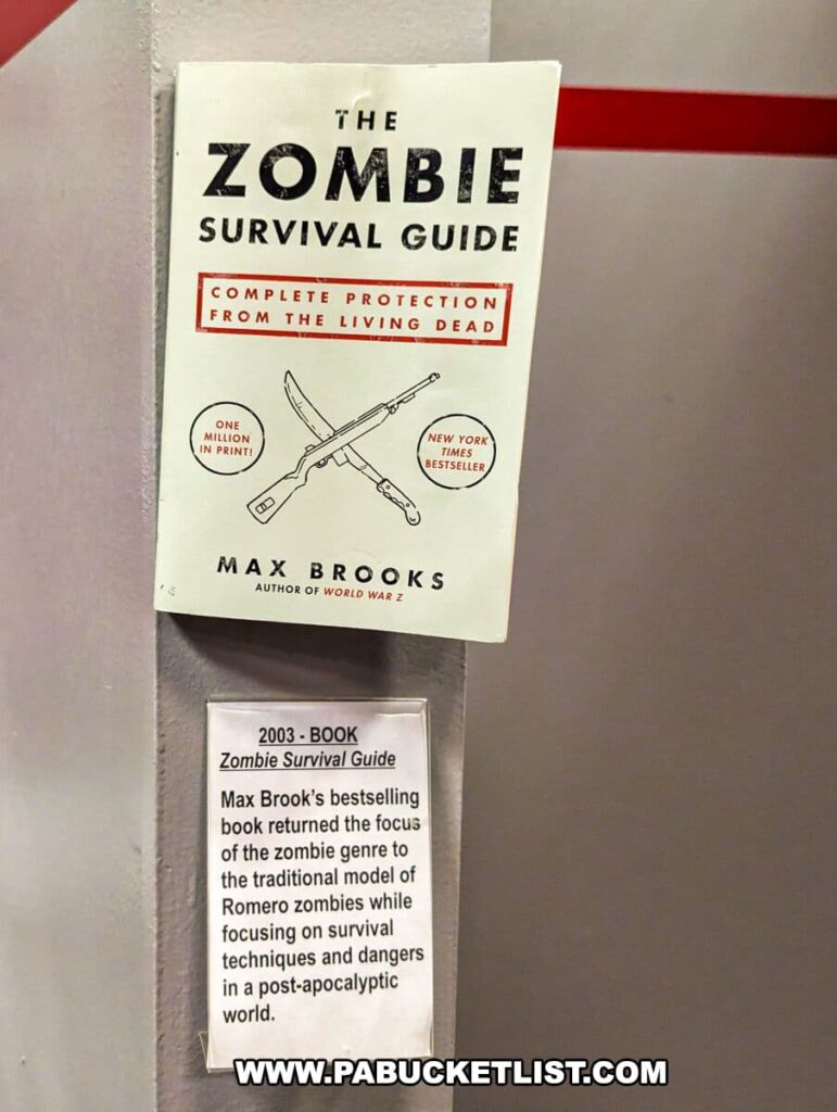 Display at the Living Dead Museum featuring 'The Zombie Survival Guide' by Max Brooks. The book cover is shown with the title and subtitle 'Complete Protection from the Living Dead' and boasts being a New York Times bestseller. Below the book, a descriptive plaque from 2003 explains how Brooks' work refocused the zombie genre on the traditional Romero model, emphasizing survival techniques and the dangers present in a post-apocalyptic world. This piece contributes to the museum's exploration of zombie culture and its impact on literature.