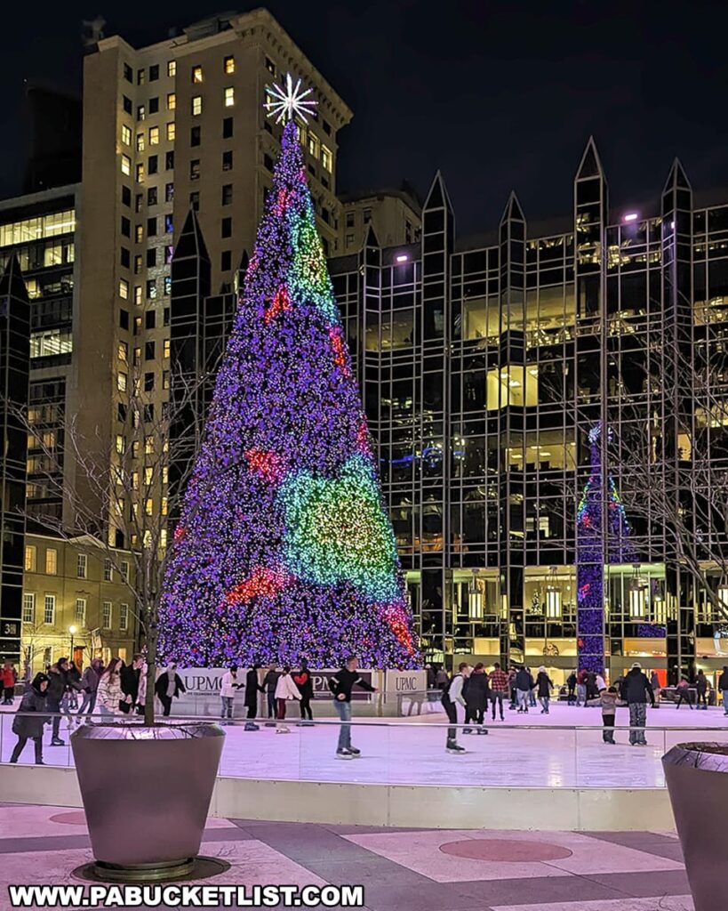 A large, brightly lit Christmas tree adorned with multicolored lights and a shining star on top stands at the center of an ice-skating rink at PPG Place in Pittsburgh, Pennsylvania. Skaters enjoy the rink, surrounded by the city's nighttime architecture and glowing windows. The festive atmosphere is underscored by the clear night sky.