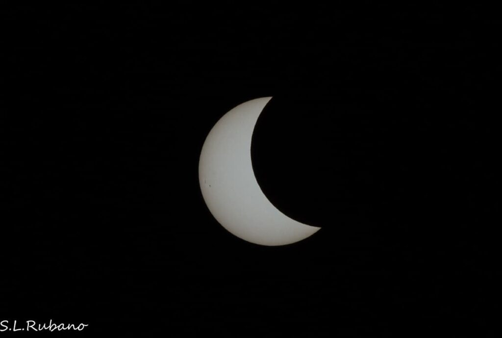 An image of a partial solar eclipse captured against a dark sky. The moon is seen covering a significant portion of the sun, leaving a bright crescent shape visible. The silhouette of the moon against the sun provides a stark contrast between the bright illuminated edge and the dark lunar surface.