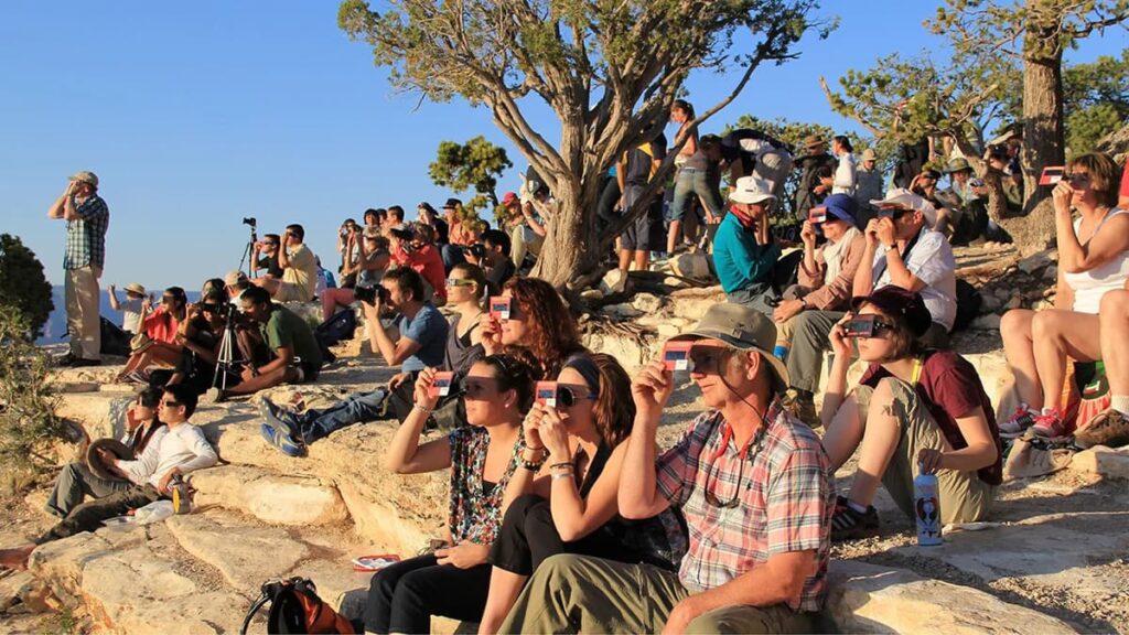 A group of people gathered outdoors on rocky terrain, preparing to view a solar eclipse. Many in the crowd are seated, looking towards the sky with eclipse glasses in place, while others are standing, some with cameras ready to capture the event. There's an air of anticipation as they wait for the celestial phenomenon. The scene is bathed in warm sunlight, suggesting it may be late afternoon. Trees and clear blue skies provide a serene backdrop to this communal viewing experience, indicative of the excitement surrounding the 2024 total solar eclipse visible in Pennsylvania.