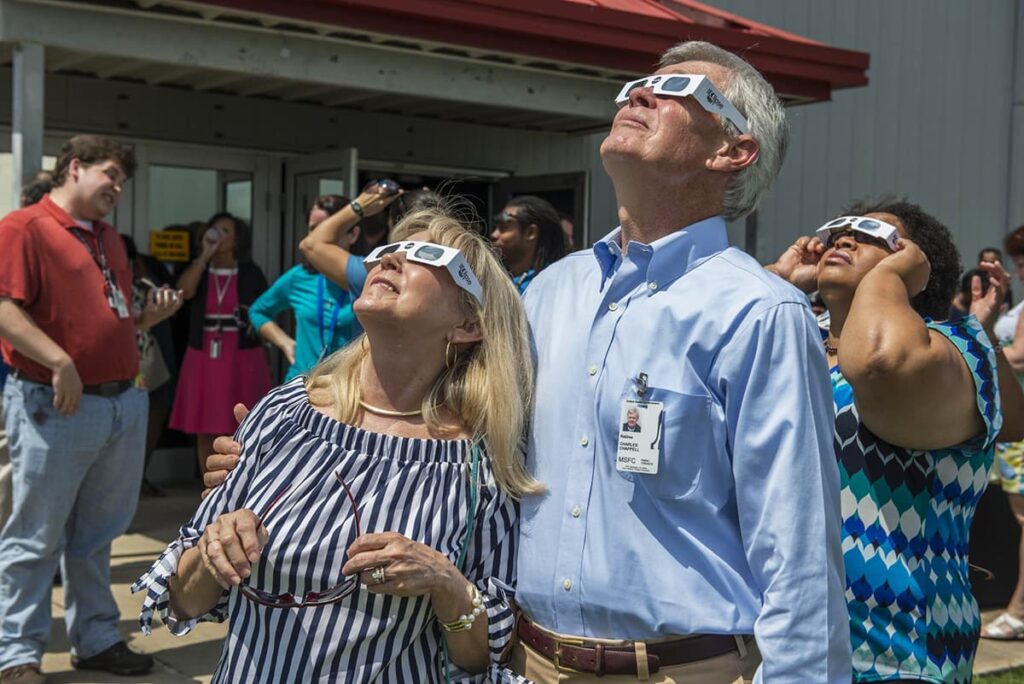 A group of people are outside on a sunny day, looking skyward with special eclipse glasses to safely view the solar eclipse. They are a mix of men and women, dressed in casual and business casual attire, indicating a diverse range of ages and backgrounds. One man in the foreground is wearing a light blue shirt with a name badge from MSFC, suggesting a connection to a scientific or technical organization. The expressions on their faces are ones of curiosity and awe as they experience the rare astronomical event.