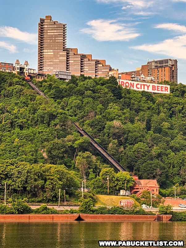 A view of the Duquesne Incline ascending Mount Washington in Pittsburgh, Pennsylvania. The incline railway is set against a lush green hillside, with the city's architecture, including a prominent multi-tiered building, in the background. A bright red "IRON CITY BEER" sign adds a splash of color to the urban landscape. The scene is captured on a sunny day with clear skies.