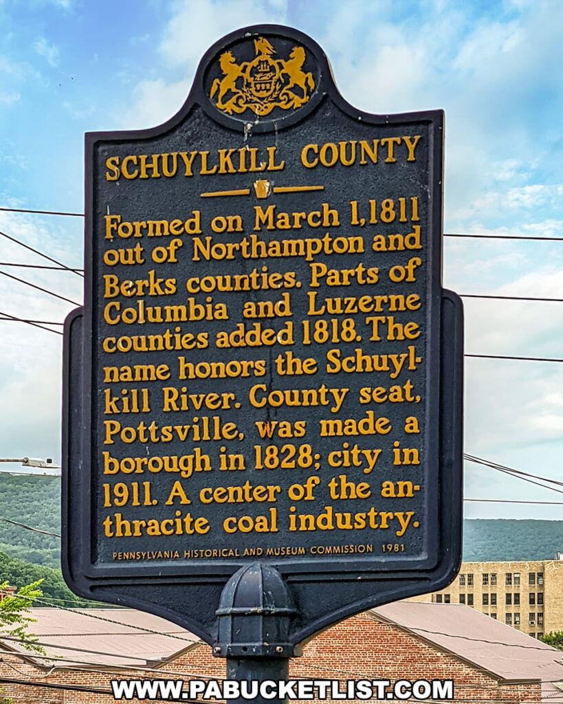 A historical marker in Pottsville, Pennsylvania, detailing the formation of Schuylkill County on March 1, 1811, from parts of Northampton and Berks counties, with additions from Columbia and Luzerne counties in 1818. The sign, which features the Pennsylvania state emblem, notes that the county's name honors the Schuylkill River and that Pottsville became the county seat in 1828 and a city in 1911. It highlights Pottsville's significance as a center of the anthracite coal industry. The sign stands on a post against a background of power lines, a hill with lush greenery, and a large building.