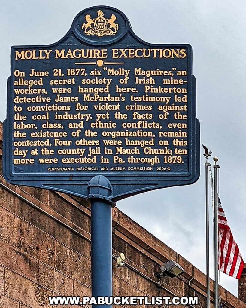 A historical marker titled 'MOLLY MAGUIRE EXECUTIONS' in Pottsville, Pennsylvania, with a detailed inscription about the hanging of six 'Molly Maguires,' an alleged secret society of Irish mine workers, on June 21, 1877. The sign references the contentious nature of the events, Pinkerton detective James McParlan's testimony, and further executions in the state through 1879. The sign, adorned with the Pennsylvania state emblem, is mounted on a blue pole against the backdrop of a brownstone wall with an American flag on a pole to the right, set under a clear sky.