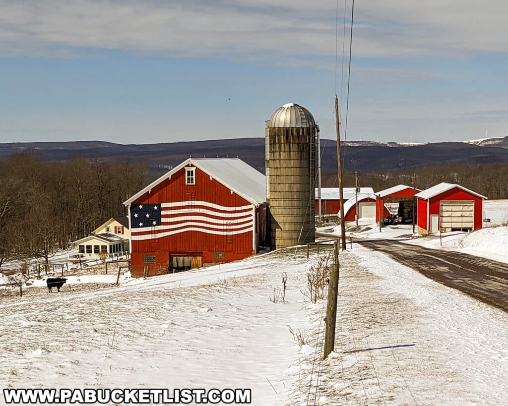 A vibrant red barn adorned with an American flag motif stands prominently on a snowy farm landscape. Near the barn, there's a traditional silo and several red outbuildings, all under a bright blue sky with distant rolling hills and wind turbines on the horizon. A black cow grazes in the foreground, and a quaint white house nestles among bare trees to the left.